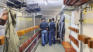 Bunkerraum mit Sitzreihen von Klappsitzen. Eine Schülergruppe verlässt gerade den Raum mit dem Rücken zum Betrachter. Im Vordergrund hängt an einer Garderobe eine beiger Parka und eine lederne Handtasche. Im Gepäckfache darüber liegt ein schwarzer Koffer.